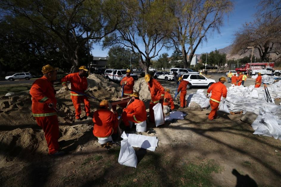 Several people wearing orange suits are shown shovelling and moving large bags.