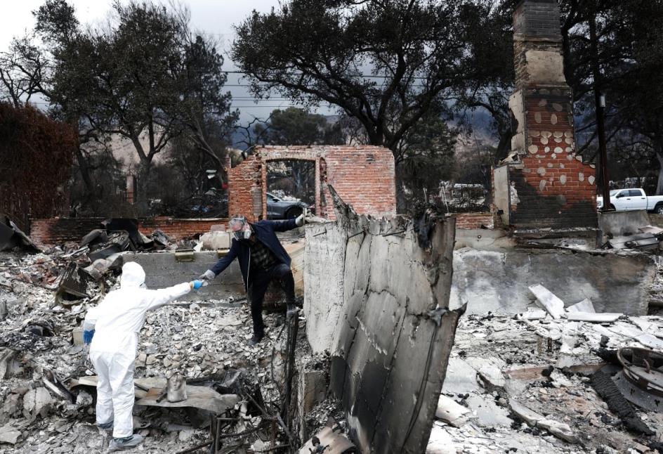 Two people are shown walking among the charred ruins of a heavily destroyed building after a fire.