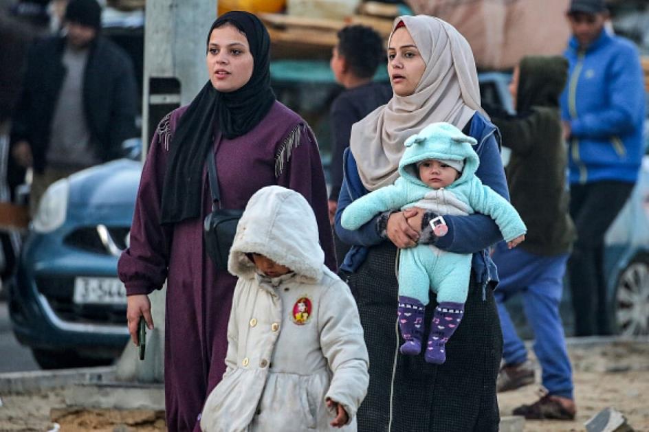 Two women walk along along a road with a young child. One carries a baby.