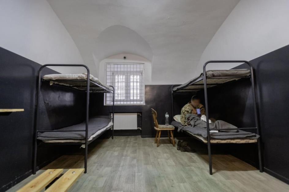 Bunk beds in a room. On the bottom left bed, a soldier is seen drinking from a cup. His face has been blurred.