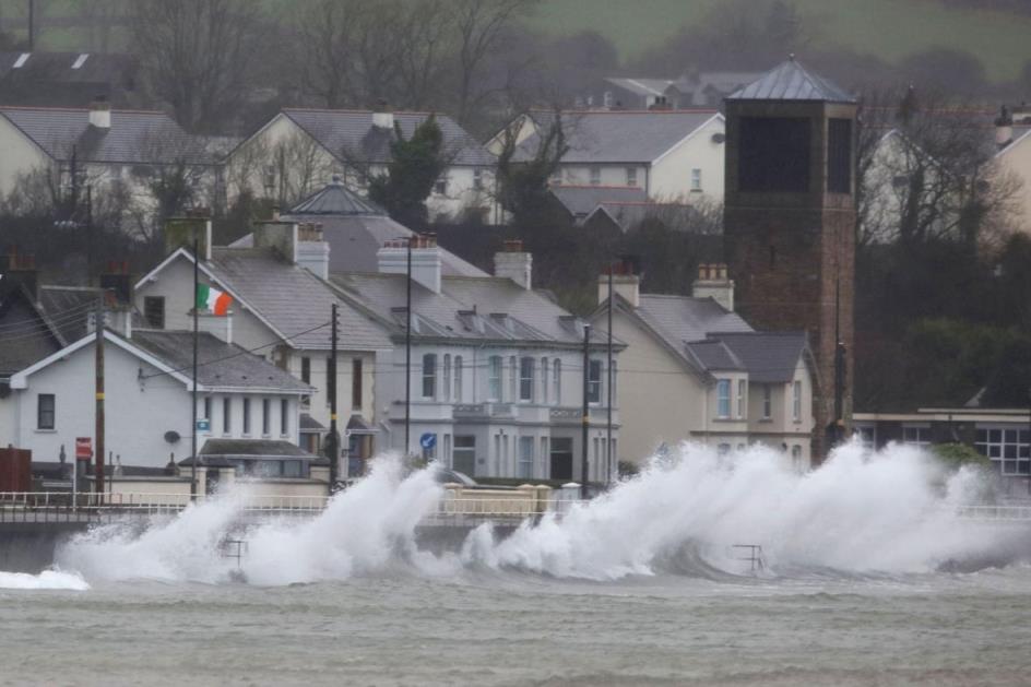 Waves from a body of water are shown high against a wall near a series of buildings.