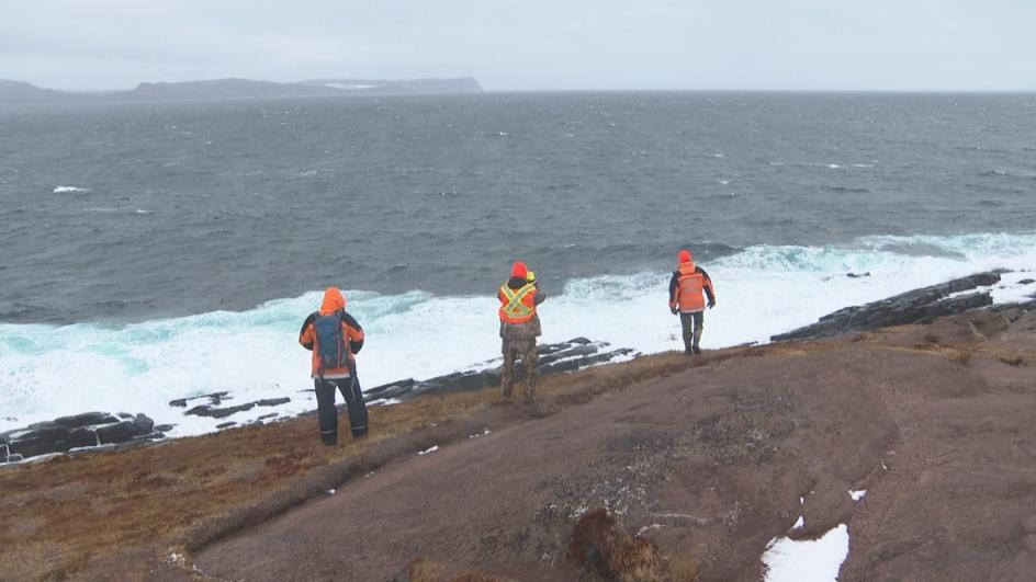 Three men wearing bright orange jackets stare into the ocean near the edge of a cliff.