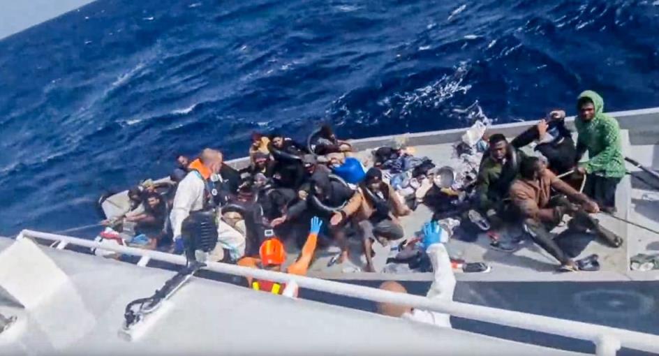 A slightly blurry photo taken from a boat looking down at a smaller boat or raft filled with people shows workers reaching out from the main boat towards the people on the smaller boat. The smaller boat is covered in clothes and other debris as well. 