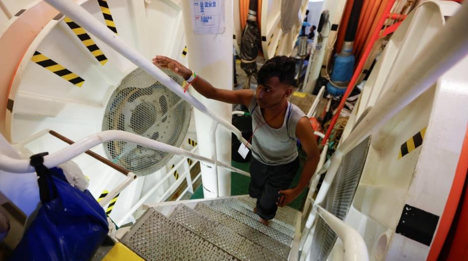 A man walks up the me<em></em>tal steps of the inside of a boat, one hand on the white railing. It is brightly lit. 