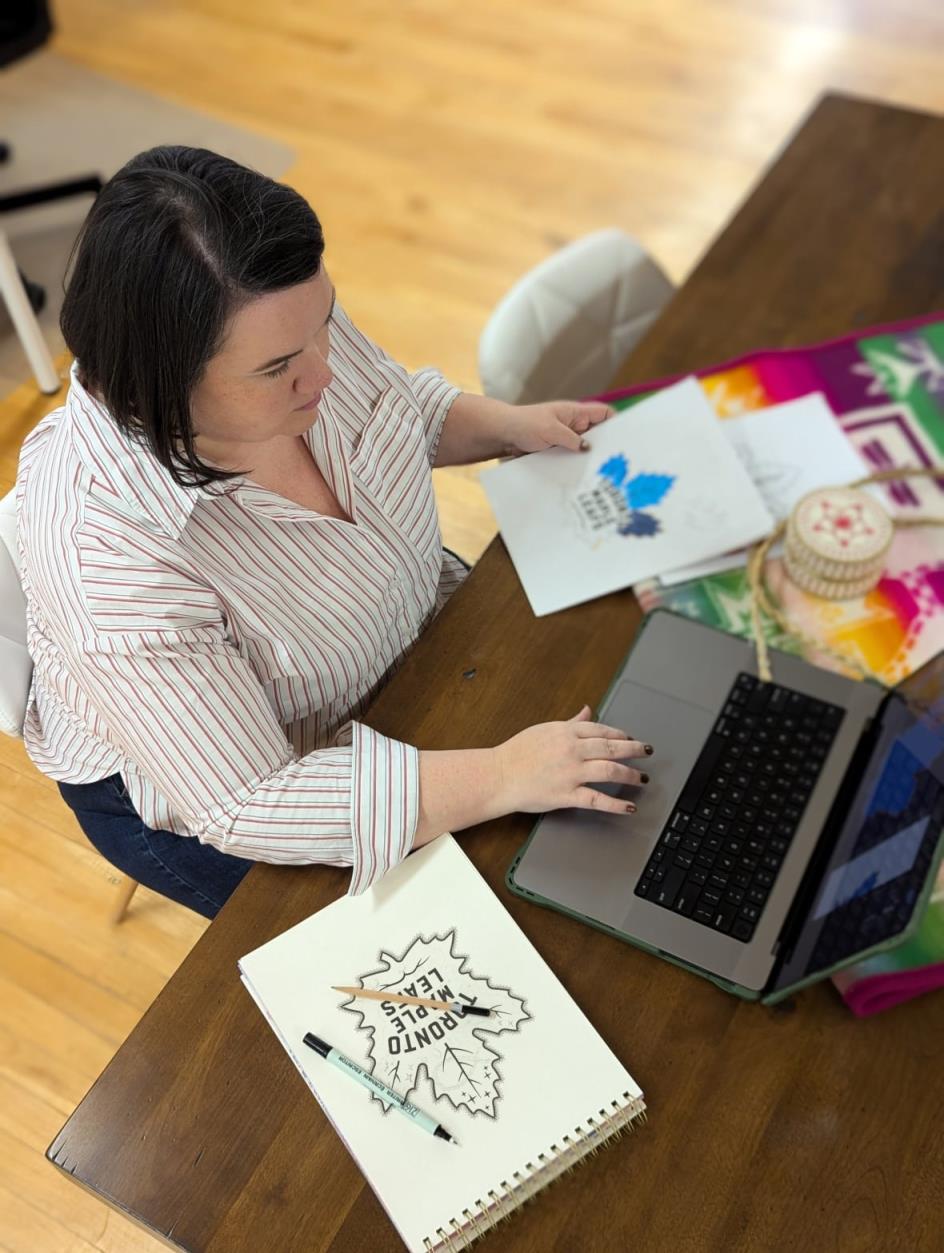A woman working on a laptop with a hand drawn image of a maple leaf on a table.